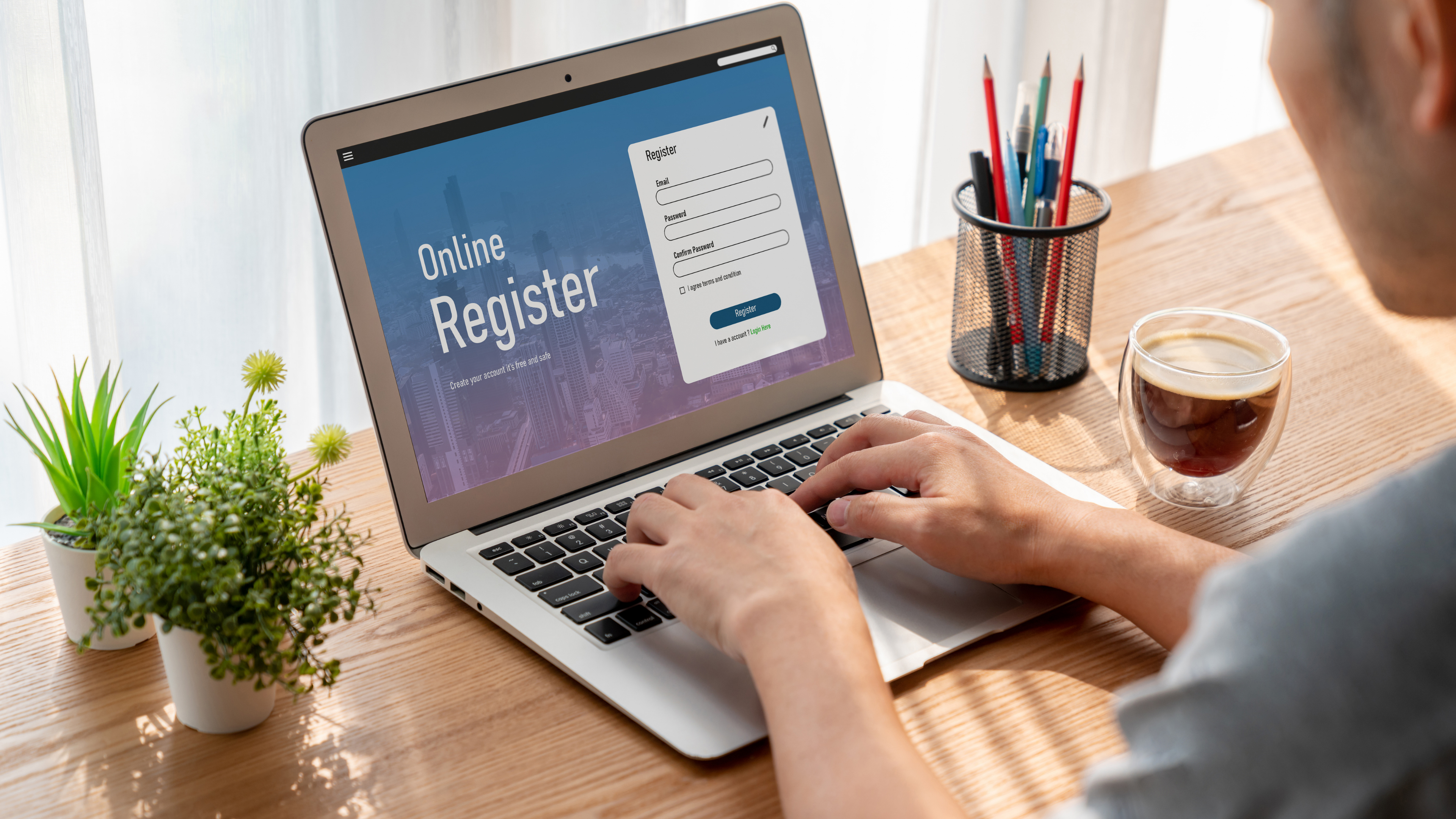 Osoba wypełnia formularz rejestracyjny online na laptopie przy biurku z kawą i roślinami.